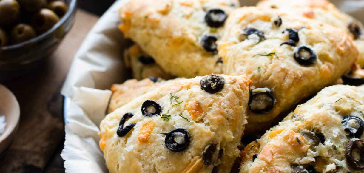 A parchment paper lined plate filled with Savory Olive Cheese Scones on a wooden table. Green and black olives in bowls in the background.
