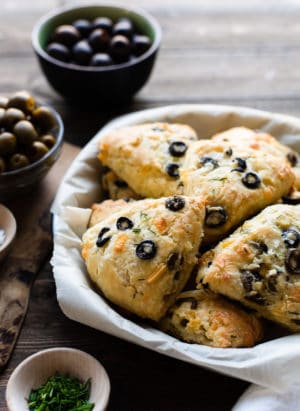 A parchment paper lined plate filled with Savory Olive Cheese Scones on a wooden table. Green and black olives in bowls in the background.