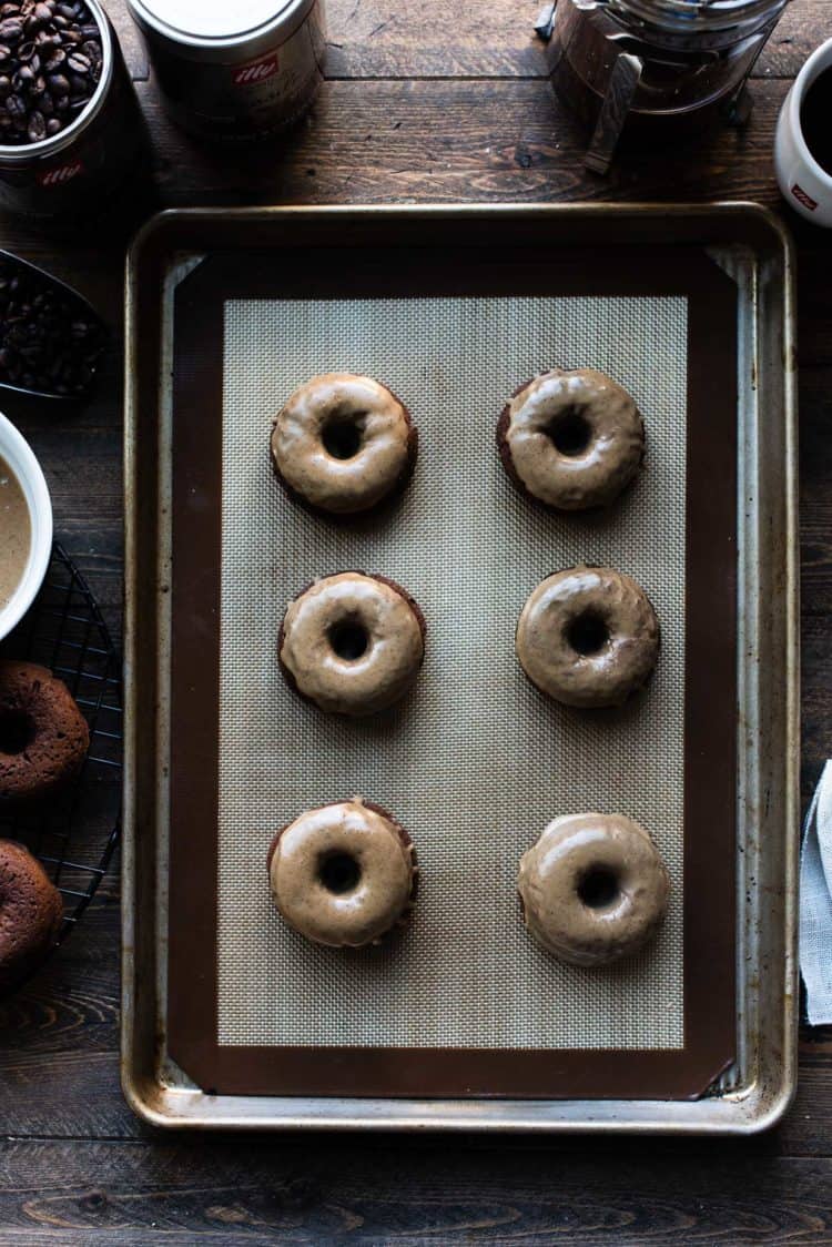 Coffee-Glazed Baked Chocolate Doughnuts on a baking tray on a wooden table with illy coffee canisters in the background.