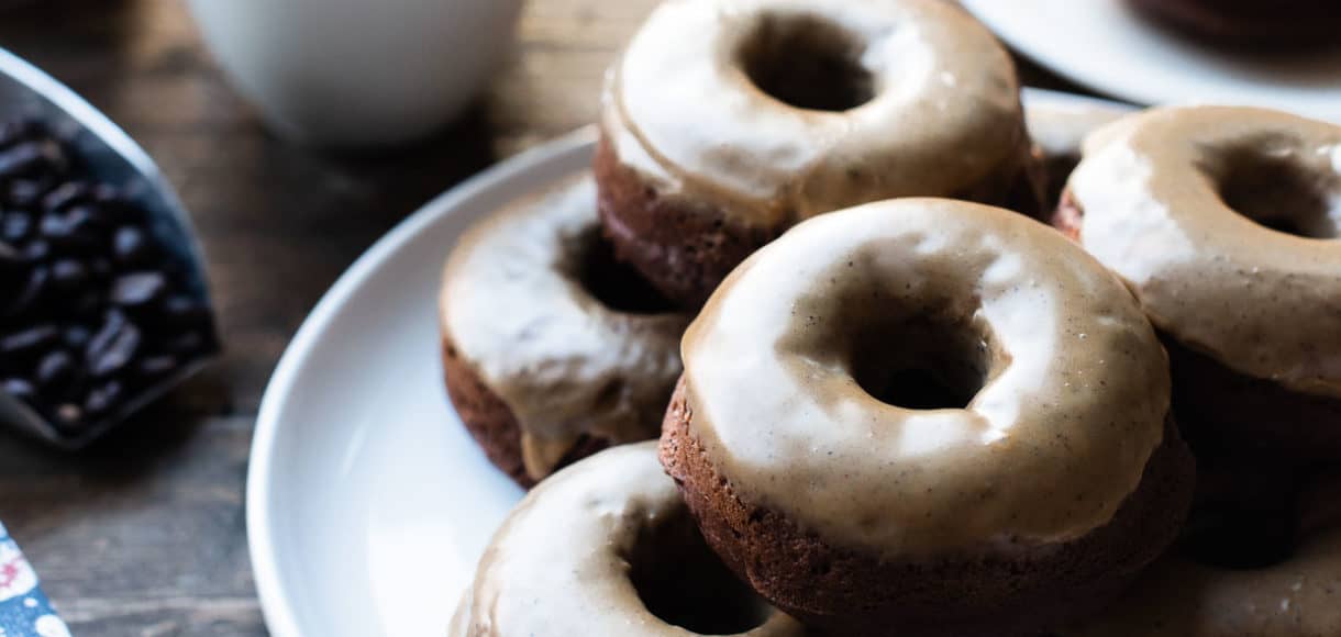 A stack of Coffee-Glazed Baked Chocolate Doughnuts on a white plate with mugs of illy coffee.