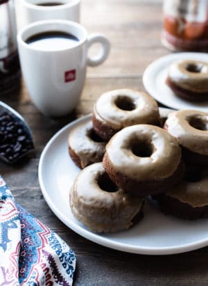 A stack of Coffee-Glazed Baked Chocolate Doughnuts on a white plate with mugs of illy coffee.