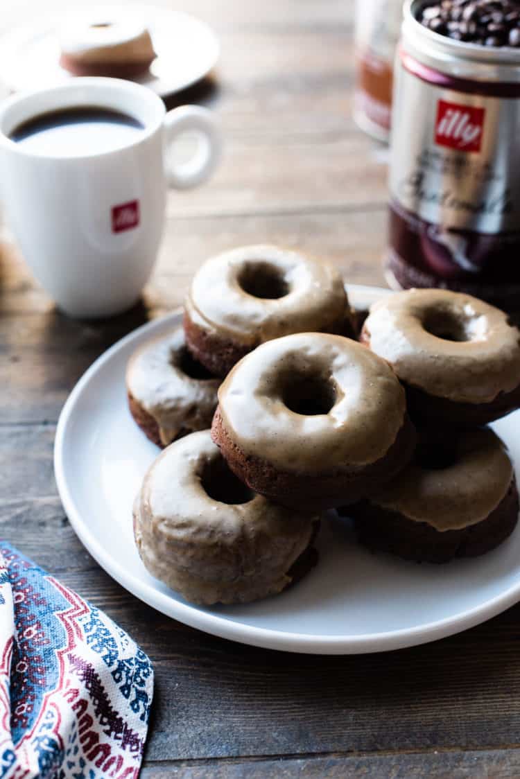 Stack of Coffee-Glazed Baked Chocolate Doughnuts on a white plate with illy coffee in the background.