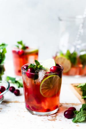 Cranberry Pomegranate Mojito in a glass garnished with limes, cranberries, pomegranate arils and mint.