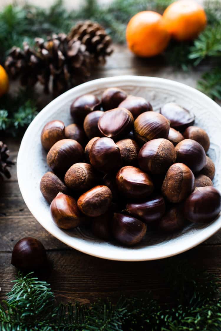 A bowl of chestnuts on a wooden table with evergreen, pinecones and oranges.