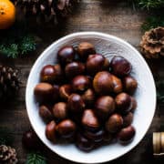 Fresh chestnuts for oven-roasted chestnut recipe in a white bowl on wooden table with evergreen, pinecones and oranges.