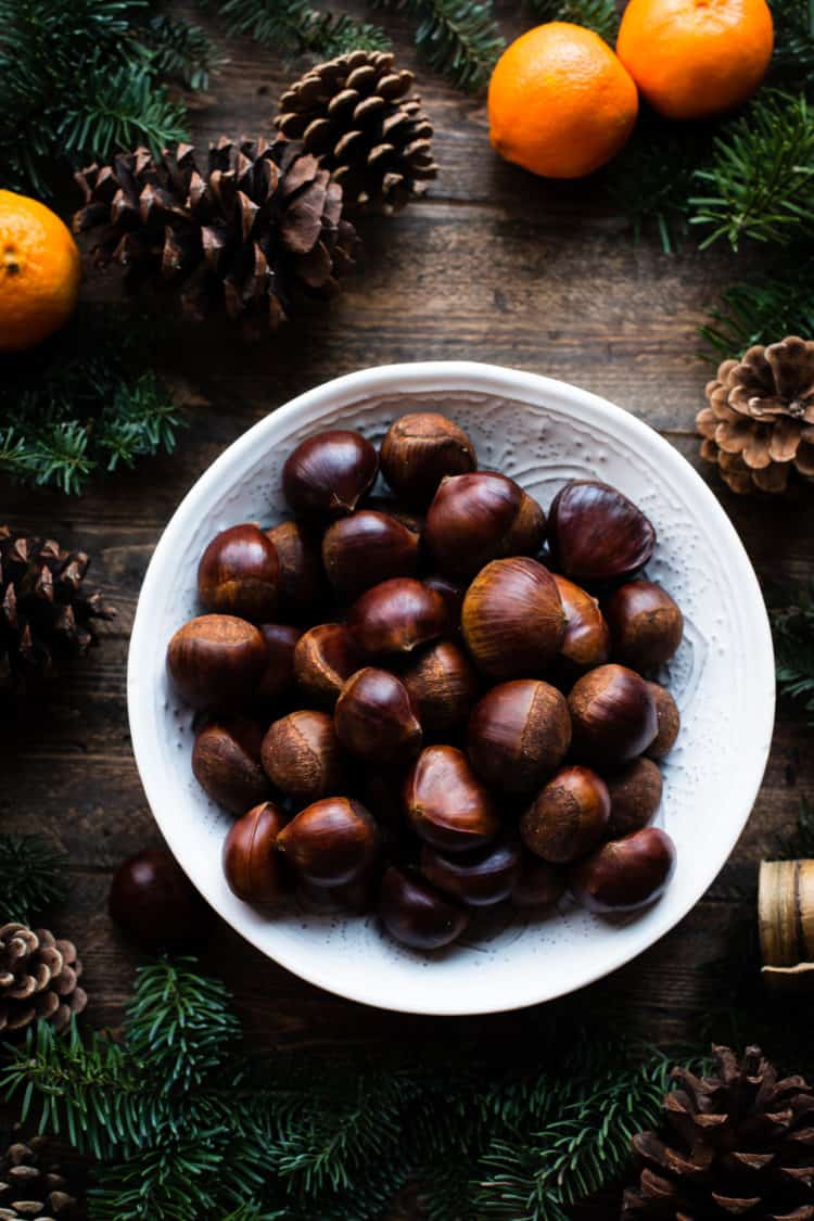 Fresh chestnuts for oven-roasted chestnut recipe in a white bowl on wooden table with evergreen, pinecones and oranges.