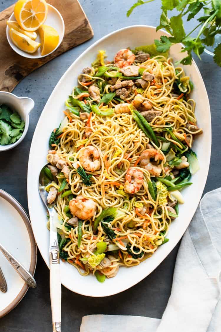 Pancit Canton on a table in a white, oval serving bowl, garnished with cilantro and lemon.