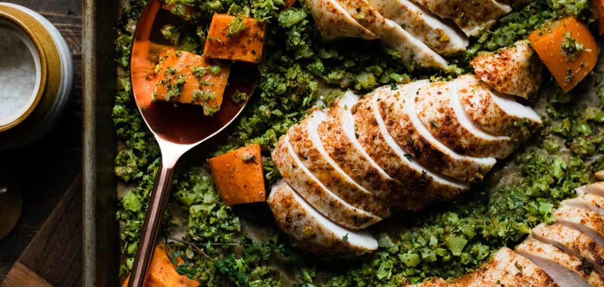Sliced chicken breast on a sheet pan with broccoli rice and sweet potatoes.