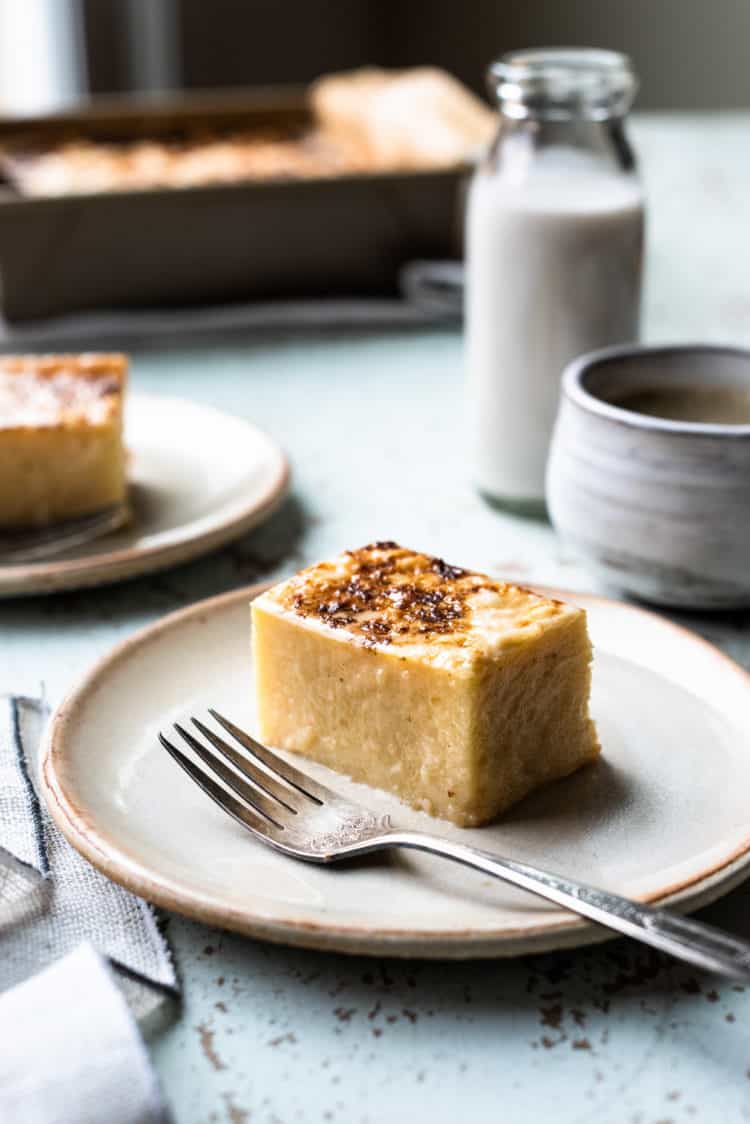 Slice of Cassava Cake on a cream plate with coffee and coconut milk in the background.