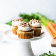 Vegan Carrot Cake Cupcakes on a small white cake stand with carrots in the background.