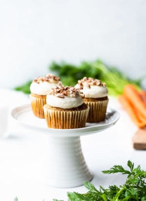 Vegan Carrot Cake Cupcakes on a small white cake stand with carrots in the background.