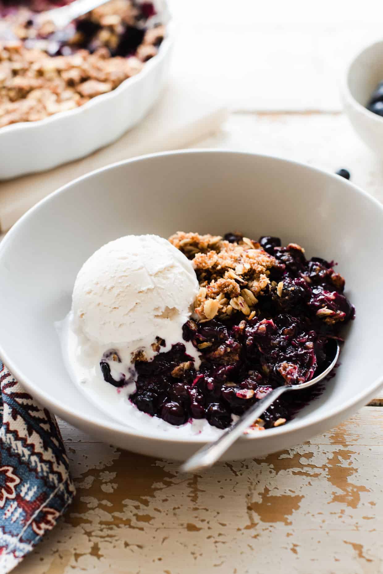 A white bowl filled with blueberry crisp and a scoop of vanilla ice cream on a rustic surface.