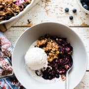 A white bowl filled with blueberry crisp and a scoop of vanilla ice cream on a rustic surface.