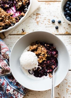A white bowl filled with blueberry crisp and a scoop of vanilla ice cream on a rustic surface.