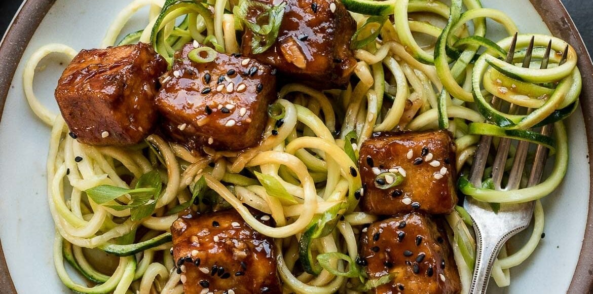 Plate of zucchini noodles (zoodles) topped with sweet and spicy crispy tofu, garnished with scallions and sesame seeds.