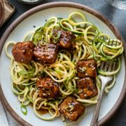 Plate of zucchini noodles (zoodles) topped with sweet and spicy crispy tofu, garnished with scallions and sesame seeds.