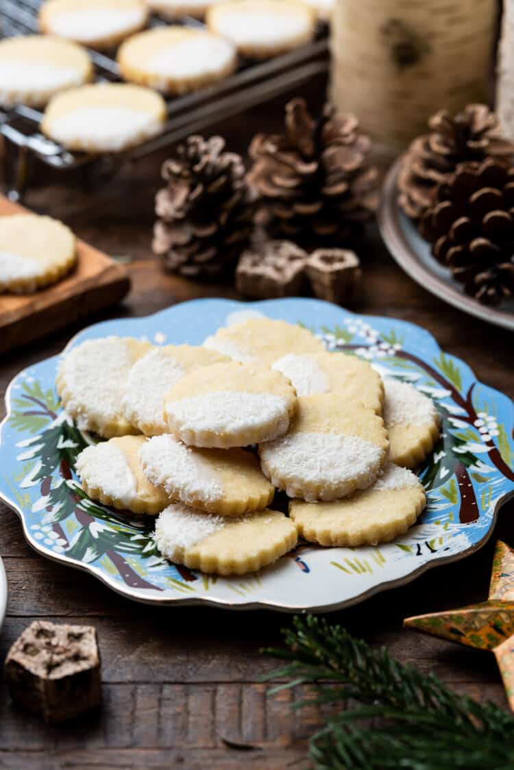 Blue holiday plate of Coconut Shortbread Cookies with coconut glaze and shredded coconut with pine cones in the background.