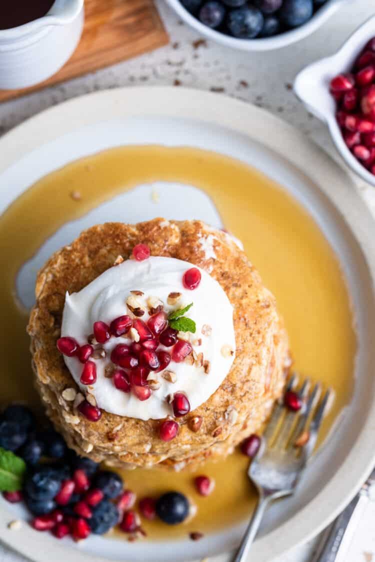 Overhead shot of oatmeal pancakes topped with coconut whipped cream, pomegranate seeds and crushed hazelnuts.