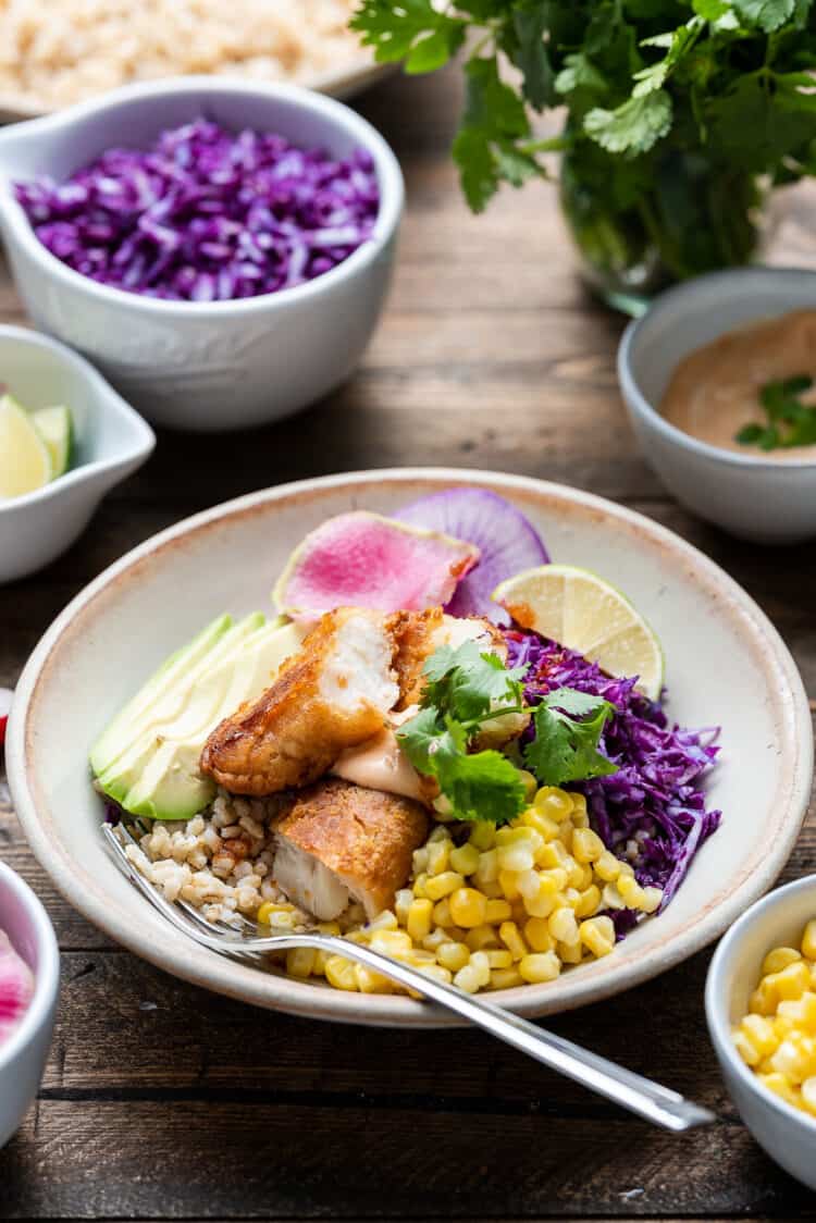 Spicy fish taco bowl with cabbage slaw in a cream bowl on a wooden table with toppings on the side.