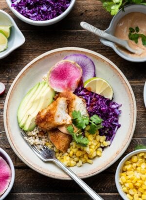 Spicy fish taco bowl with cabbage slaw in a cream bowl on a wooden table with toppings on the side.