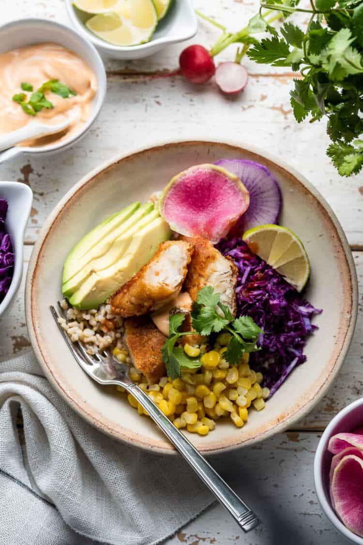 Spicy fish taco bowl with cabbage slaw in a cream bowl on a white table with toppings on the side.