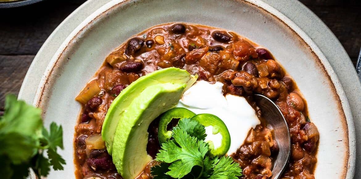 A bowl of vegan chili with beans, avocado and cilantro and a side of corn bread.