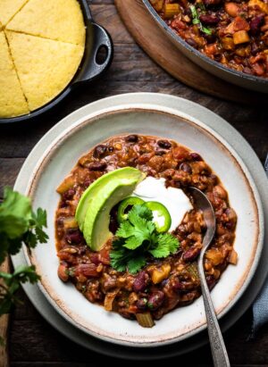 A bowl of vegan chili with beans, avocado and cilantro and a side of corn bread.
