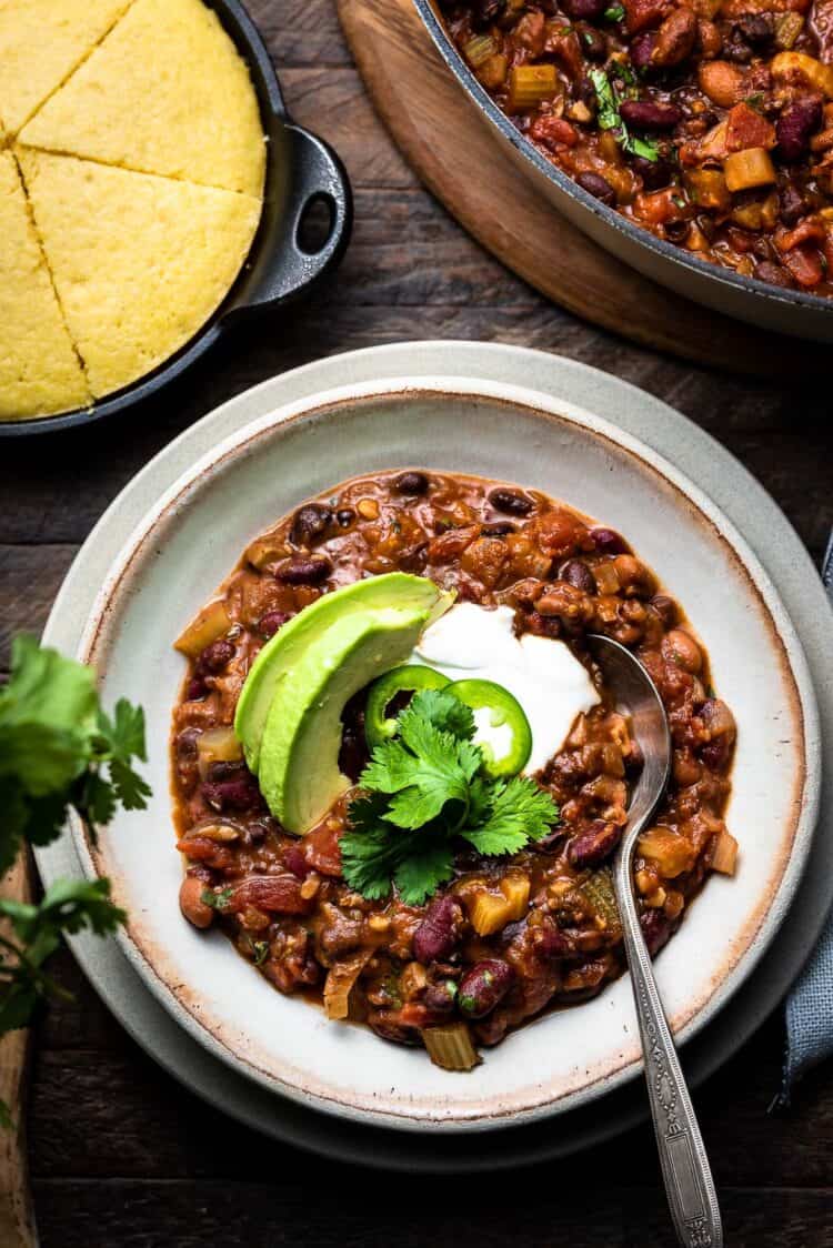 A bowl of vegan chili with beans, avocado and cilantro and a side of corn bread.