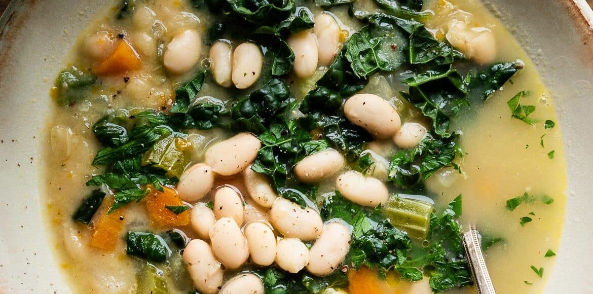 White bean soup with kale, spinach and veggies in a cream-colored bowl and croutons on the side.