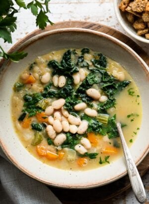 White bean soup with kale, spinach and veggies in a cream-colored bowl and croutons on the side.