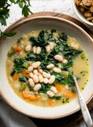 White bean soup with kale, spinach and veggies in a cream-colored bowl and croutons on the side.