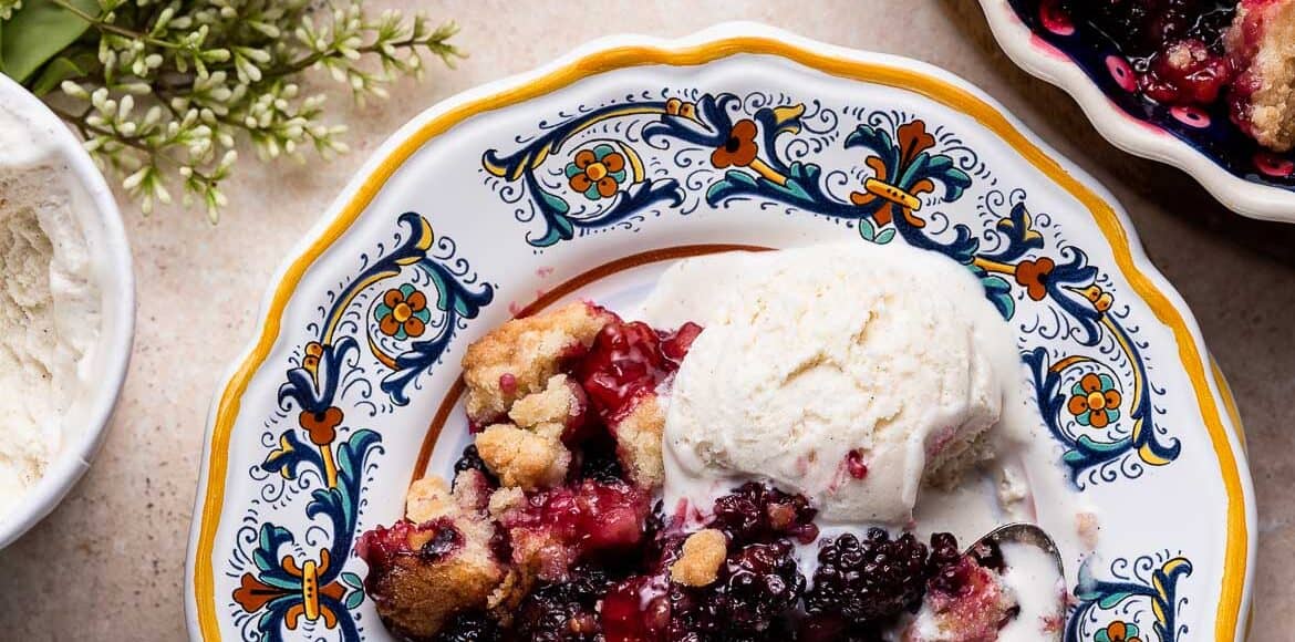 Blackberry Crumble with vanilla ice cream served on a yellow edged plate with a spoon.