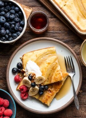 Two sheet pan banana pancakes on a white plate topped with cream, bananas, berries and maple syrup on a wooden table.