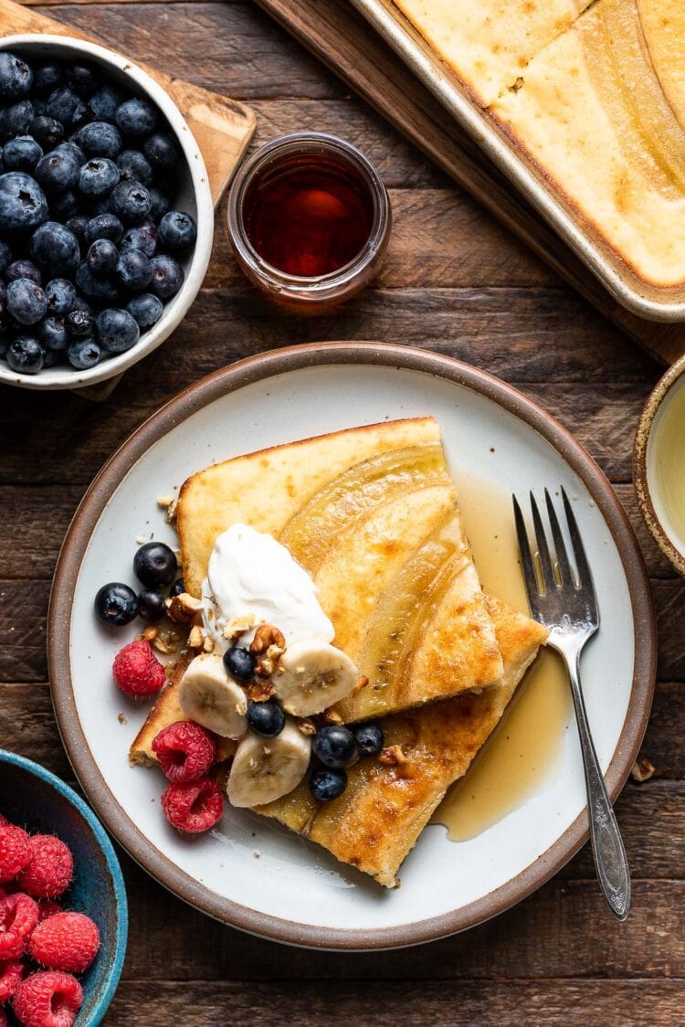 Two sheet pan banana pancakes on a white plate topped with cream, bananas, berries and maple syrup on a wooden table.