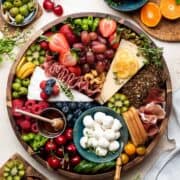 A cheese and charcuterie board with fruits, berries, vegetables and crackers in a wooden platter on a cream table surrounded by ingredients.