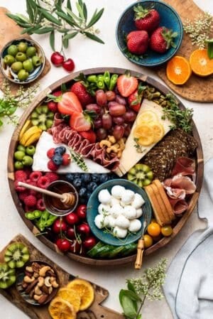 A cheese and charcuterie board with fruits, berries, vegetables and crackers in a wooden platter on a cream table surrounded by ingredients.