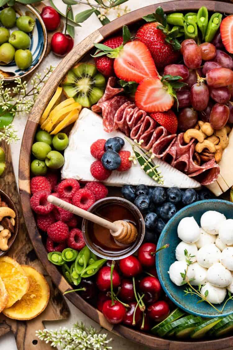 Close up of a cheese charcuterie board in a wooden platter with fresh fruits, vegetables, olives.