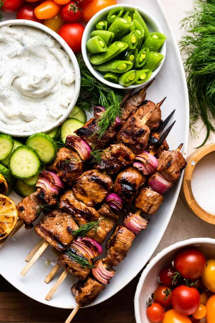 Grilled Pork Skewers made with Balsamic Marinade on a white serving platter with tzatziki and fresh vegetables.