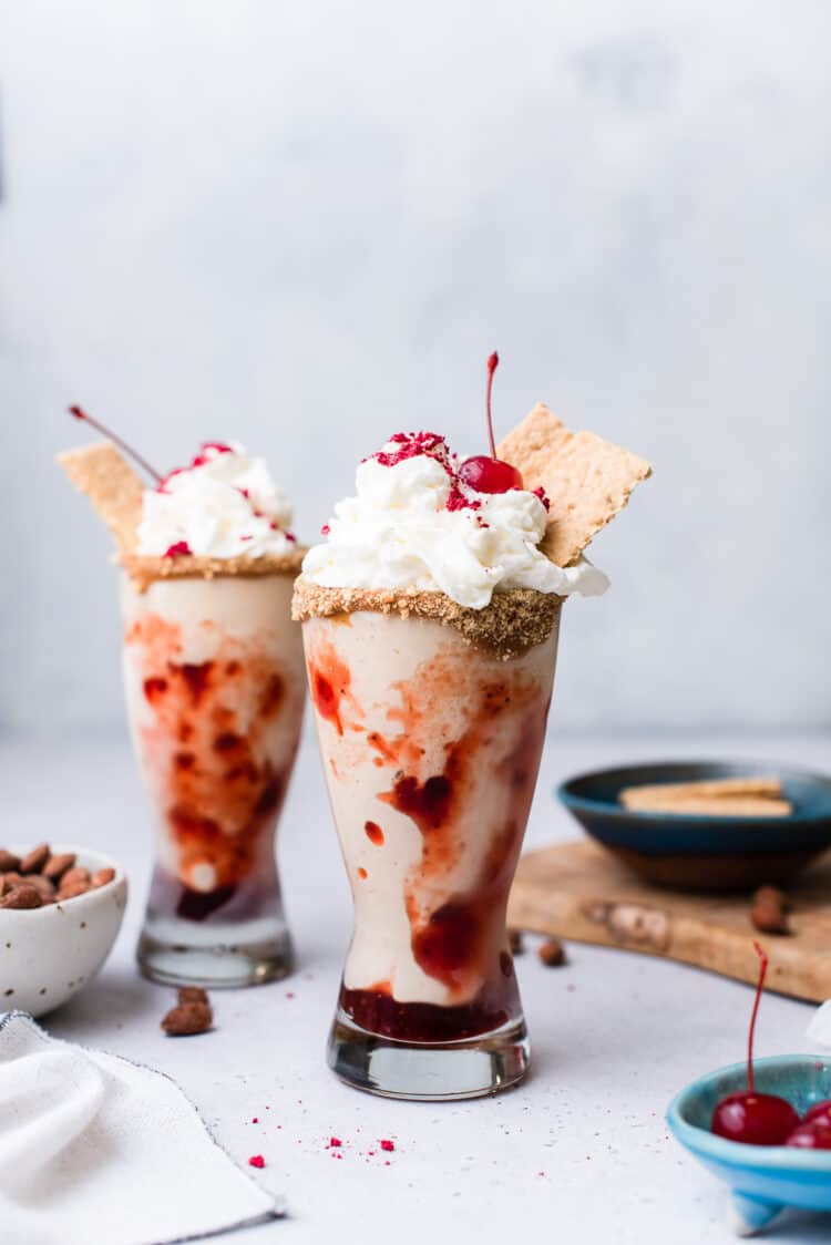 Almond Butter and Jelly Vegan Milkshake in a glass topped with non-dairy whipped cream, graham crackers and a maraschino cherry.