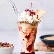 Almond Butter and Jelly Vegan Milkshake in a glass topped with non-dairy whipped cream, graham crackers and a maraschino cherry.