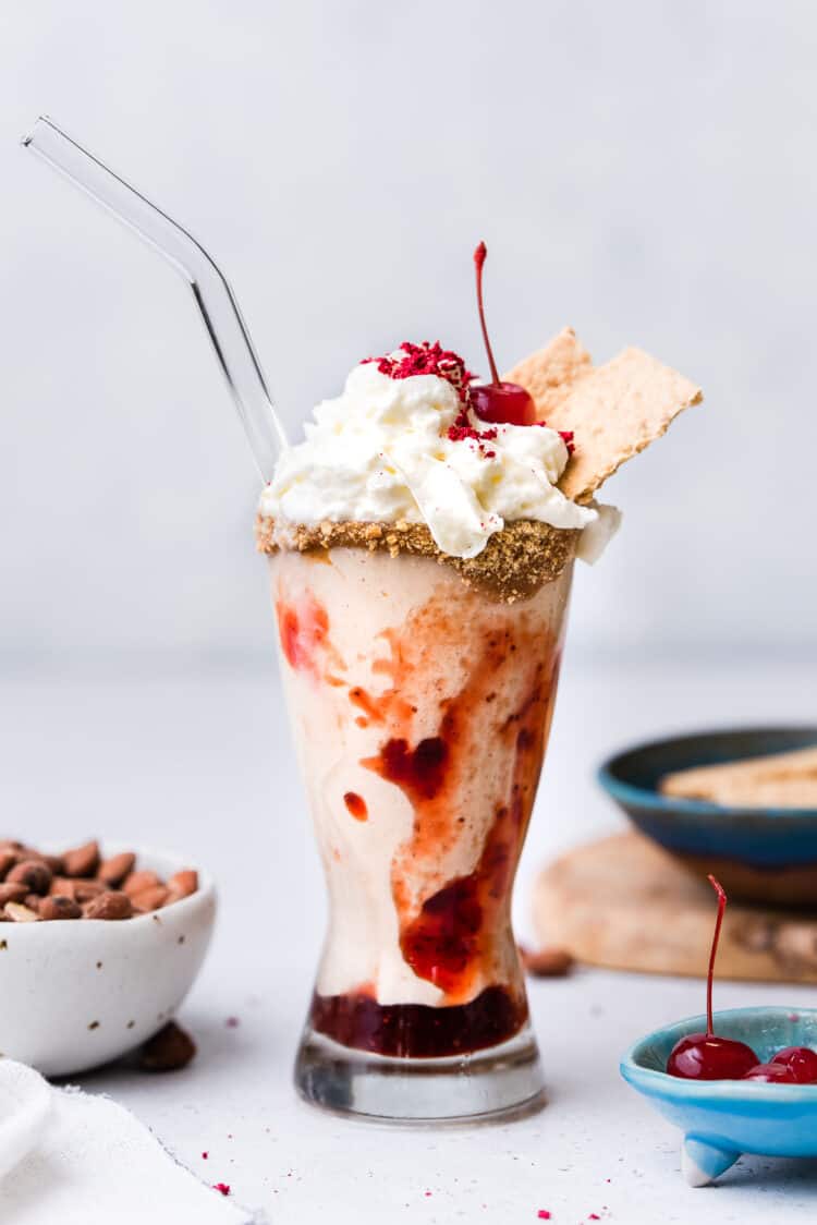 Almond Butter and Jelly Vegan Milkshake in a glass topped with non-dairy whipped cream, graham crackers and a maraschino cherry.