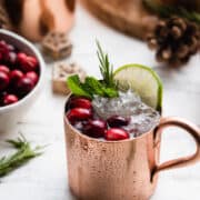Pomegranate Cranberry Moscow Mule in a copper cup garnished with cranberries, mint, lime and rosemary.
