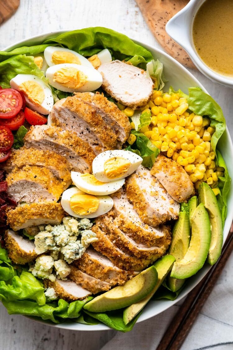 Crispy chicken Cobb salad in a white round bowl with air fryer breaded chicken, hard boiled eggs, avocado, tomato , bacon, corn, blue cheese and lettuce with Cobb salad dressing on the side.