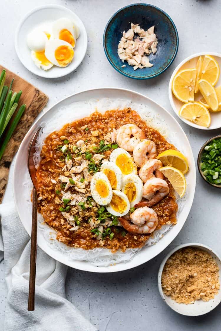 Pancit Palabok in a white bowl garnished with shrimp and hard boiled eggs and garnishes in bowl.