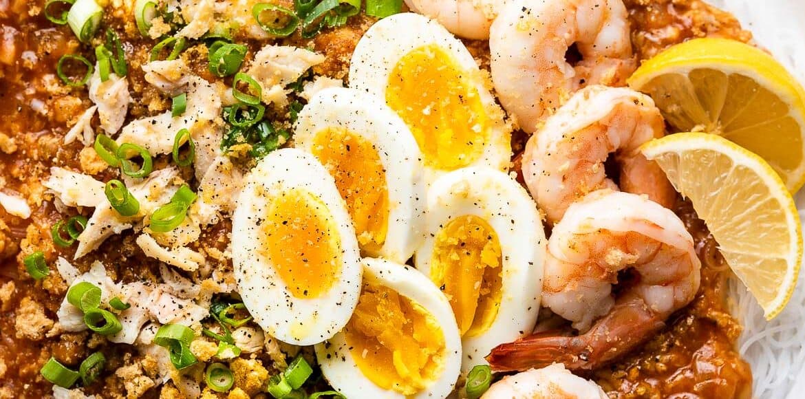 Pancit Palabok in a white bowl garnished with shrimp and hard boiled eggs.