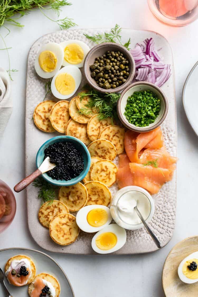 Caviar and blini board with smoked salmon, creme fraiche, chives, capers, and hardboiled eggs on a platter.