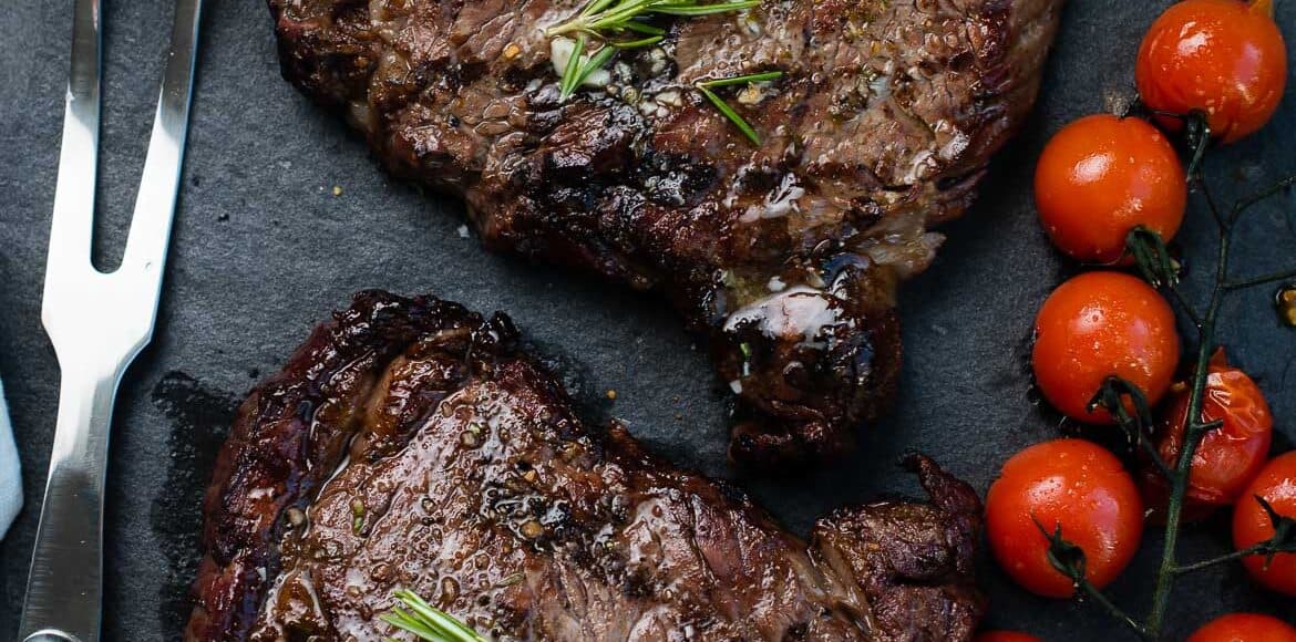 Grilled dry aged rib eye steaks resting on a slate platter with grilled tomatoes, garlic, and garlic butter.