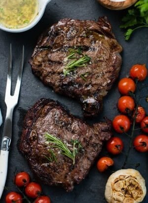 Grilled dry aged rib eye steaks resting on a slate platter with grilled tomatoes, garlic, and garlic butter.