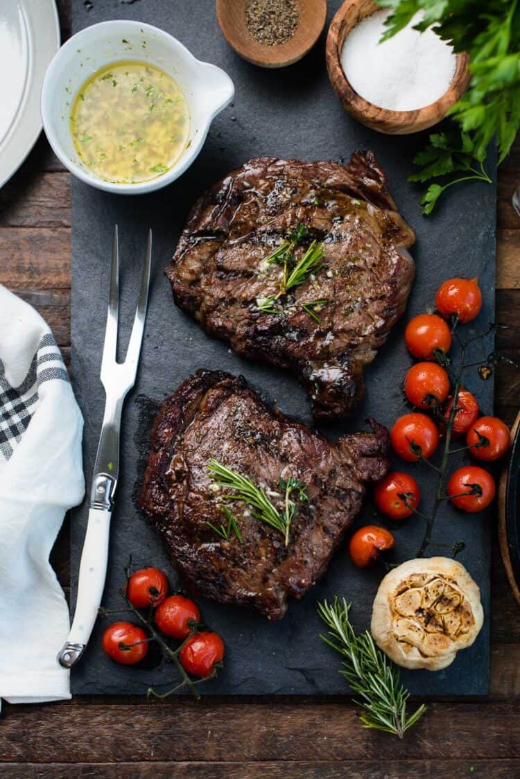 Grilled dry aged rib eye steaks resting on a slate platter with grilled tomatoes, garlic, and garlic butter.