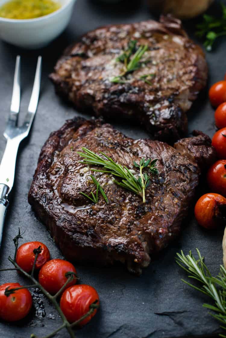 Grilled dry aged rib eye steaks resting on a slate platter with grilled tomatoes, garlic, and garlic butter.